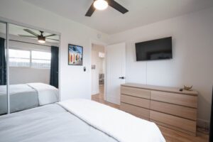 Modern bedroom with white walls, a ceiling fan, and wall-mounted TV above a wooden dresser.