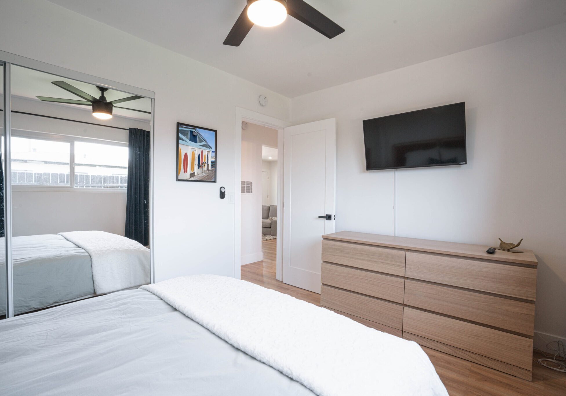 Modern bedroom with white walls, a ceiling fan, and wall-mounted TV above a wooden dresser.